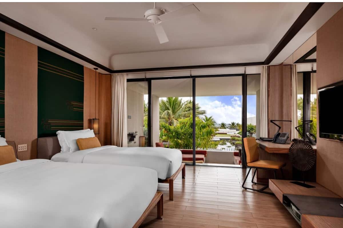 A hotel room with two beds, a desk with a chair, a TV, and a view of palm trees through large windows.