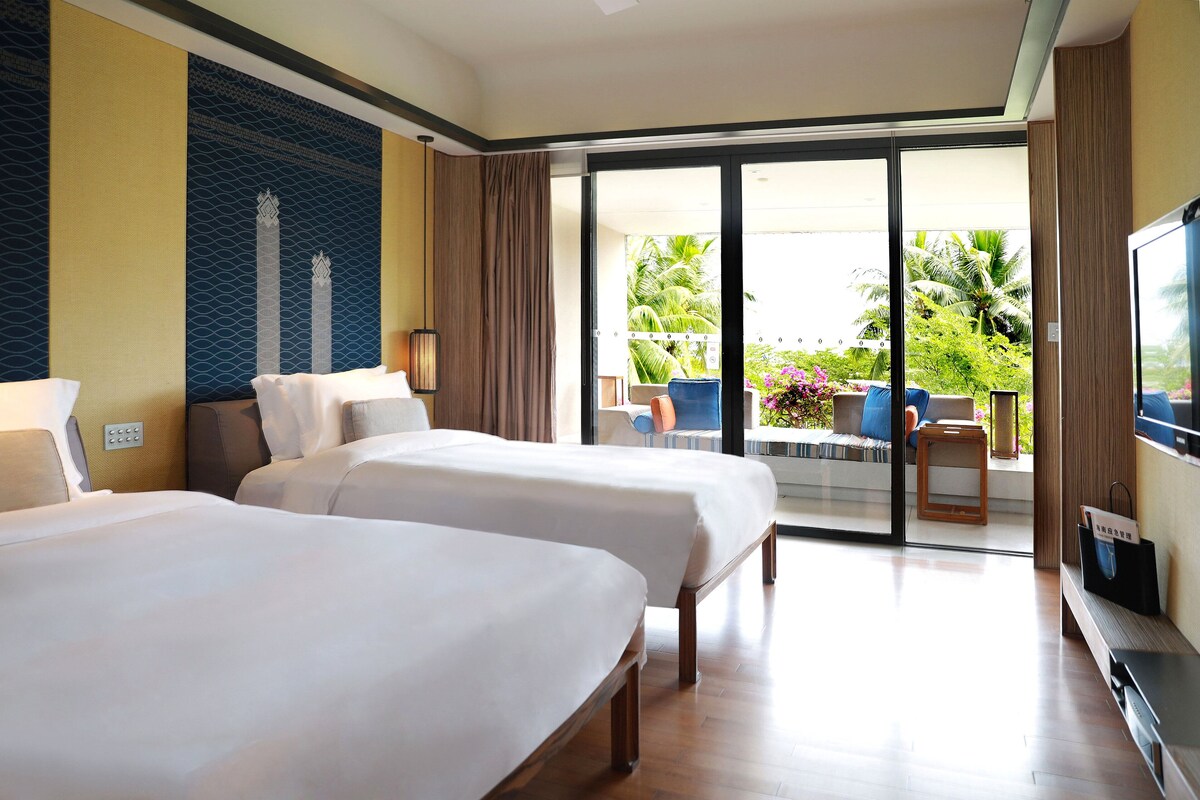 A hotel room with two beds, a TV, a balcony with a view of palm trees and a pool.
