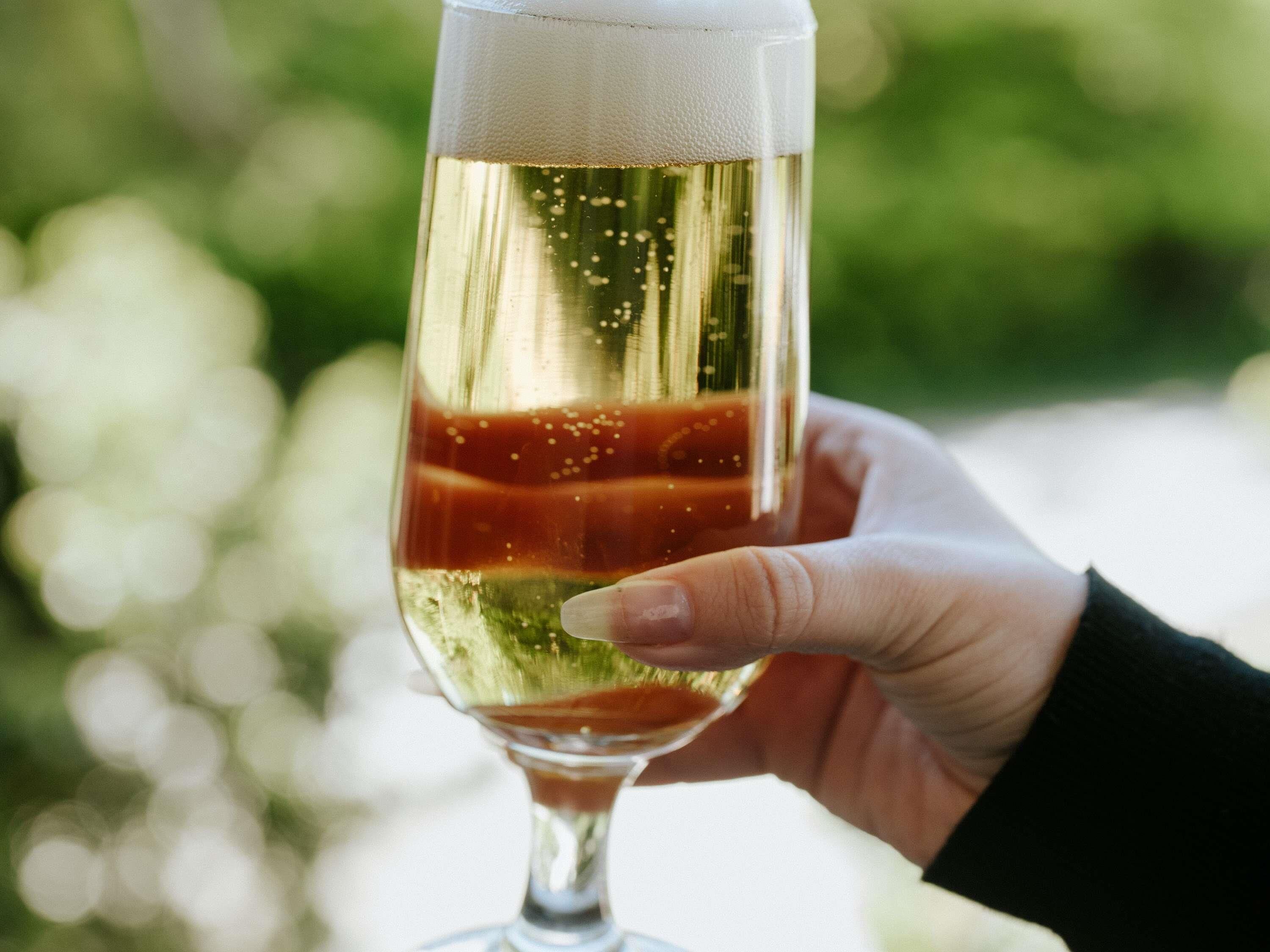 A hand holding a glass with a layered beverage, possibly champagne, with a blurred green background.