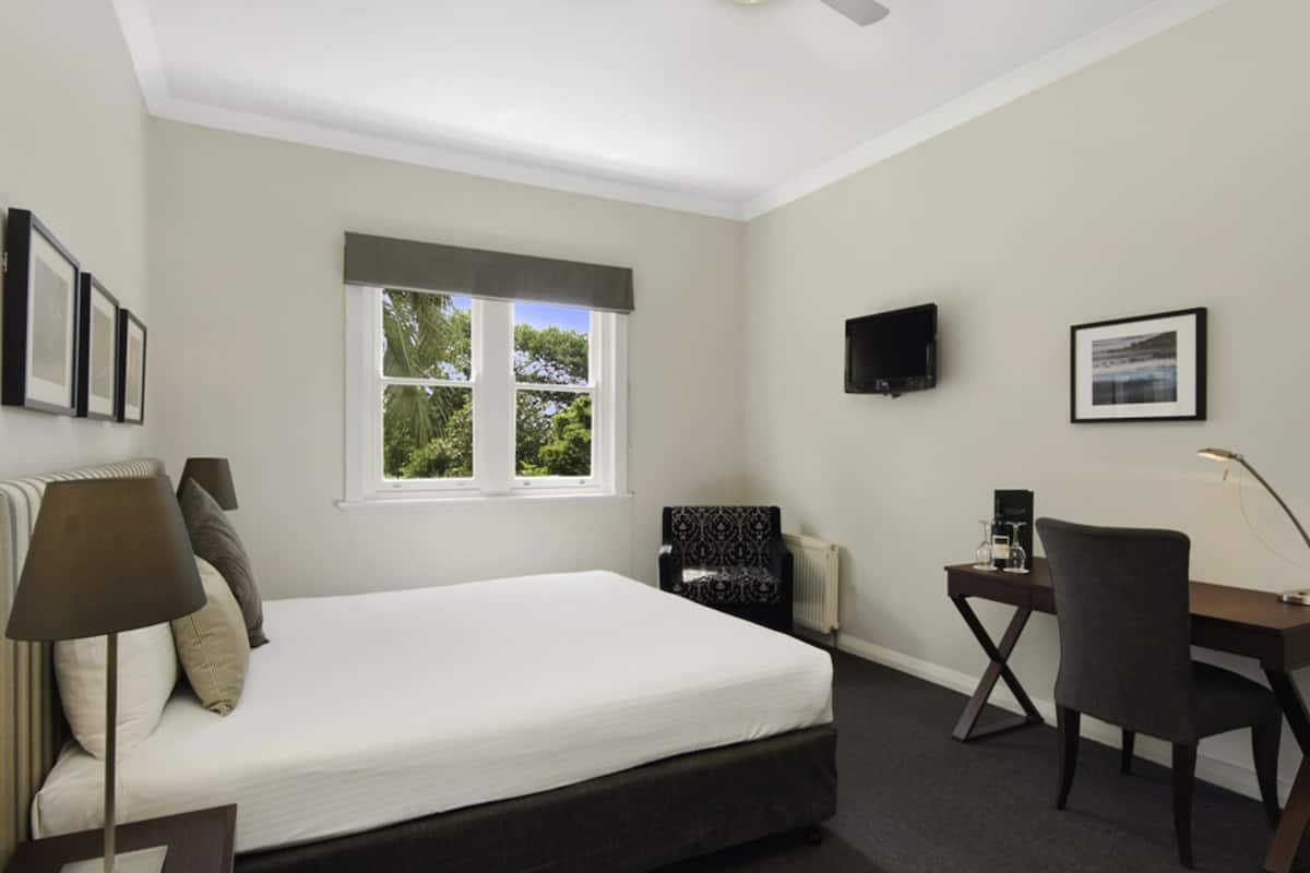 A bedroom with a bed, a desk, a chair, a television, and a window with a view of trees.