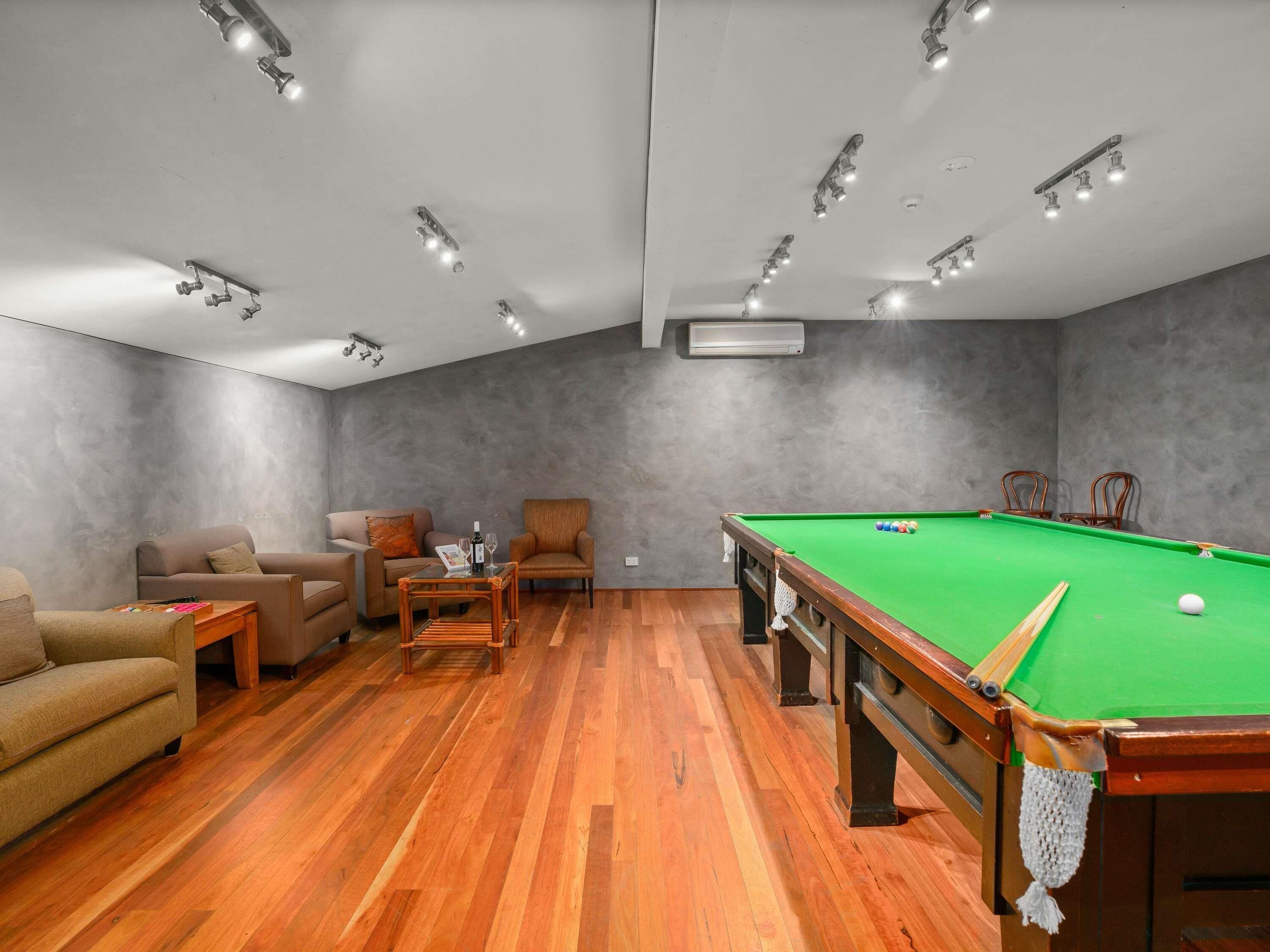 A room with a pool table, sofas, a small table with a bottle and glasses, and recessed ceiling lights.