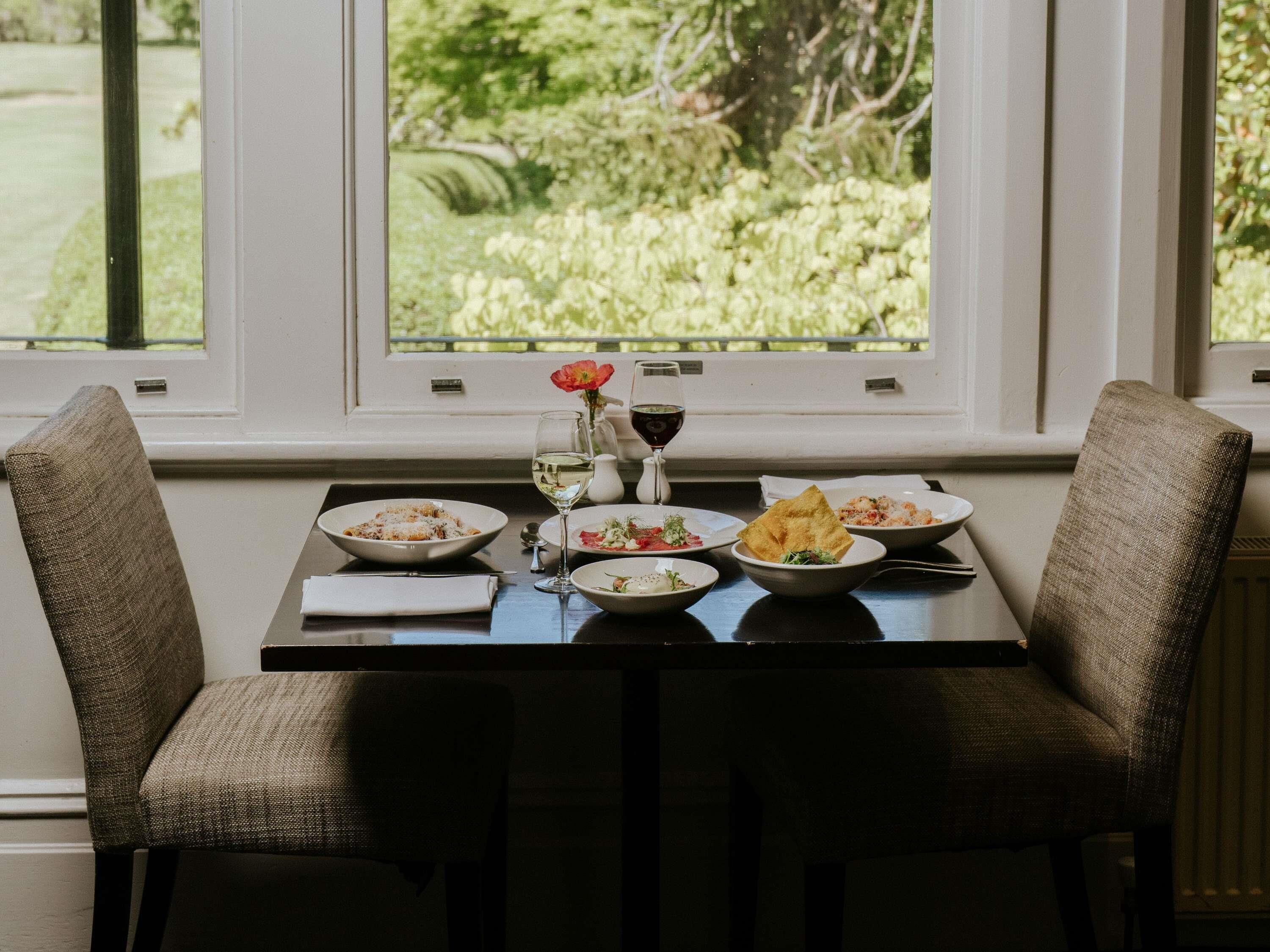 A dining table with four plates of food, a wine glass, and a vase with a flower. Two upholstered chairs are positioned on either side of the table.