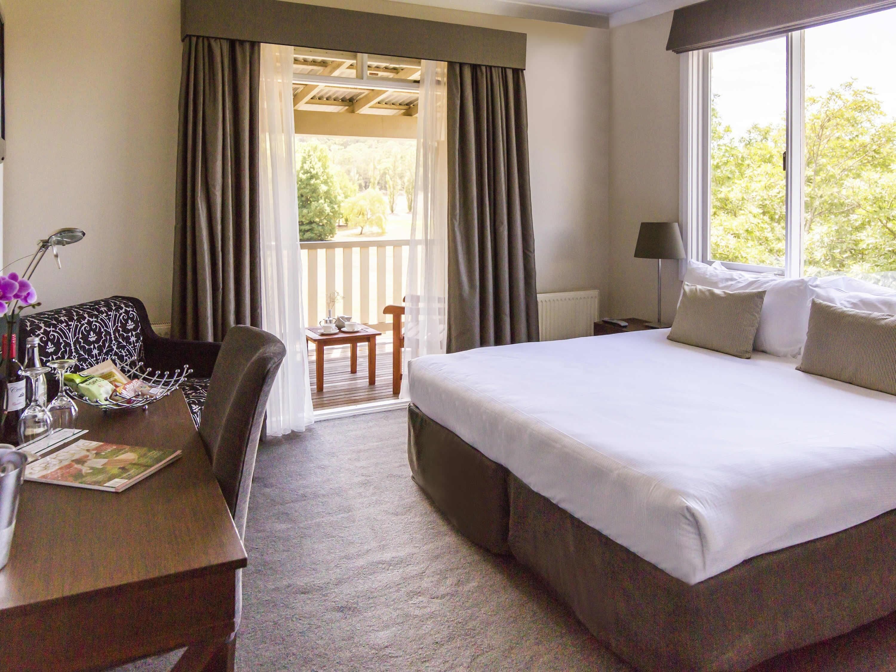 Une chambre d’hôtel comprenant un lit, un bureau avec une chaise, une petite table avec une chaise, une fenêtre avec des rideaux et une vue sur la verdure à l’extérieur.
