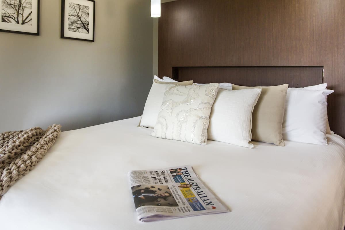 A neatly made bed with white bedding and a folded newspaper on it. There are two framed pictures on the wall above the bed.