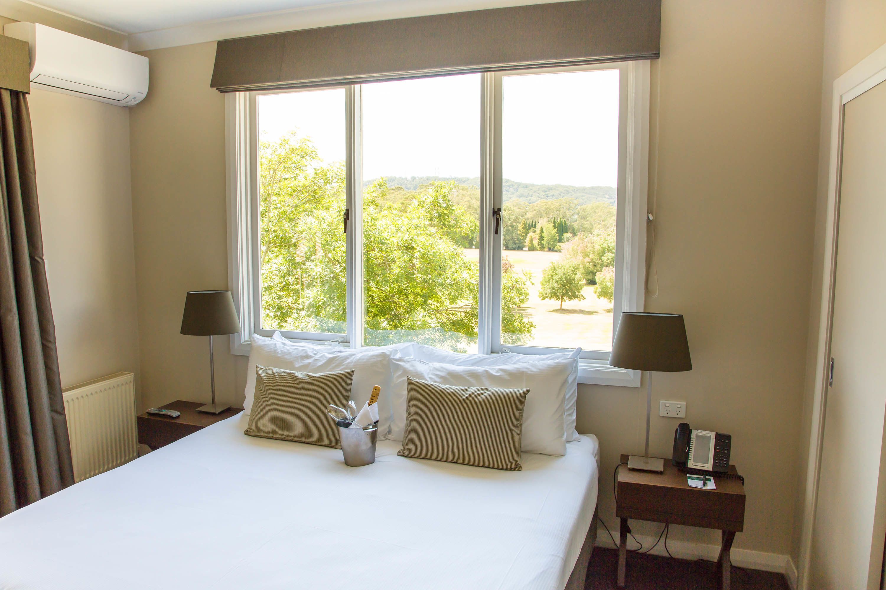 A hotel room with a large bed, two bedside lamps, a small table with a phone and a lamp, and a window with a view of trees and hills.