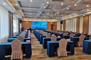 A conference room with rows of tables and chairs, a large screen at the front, and a carpeted floor.