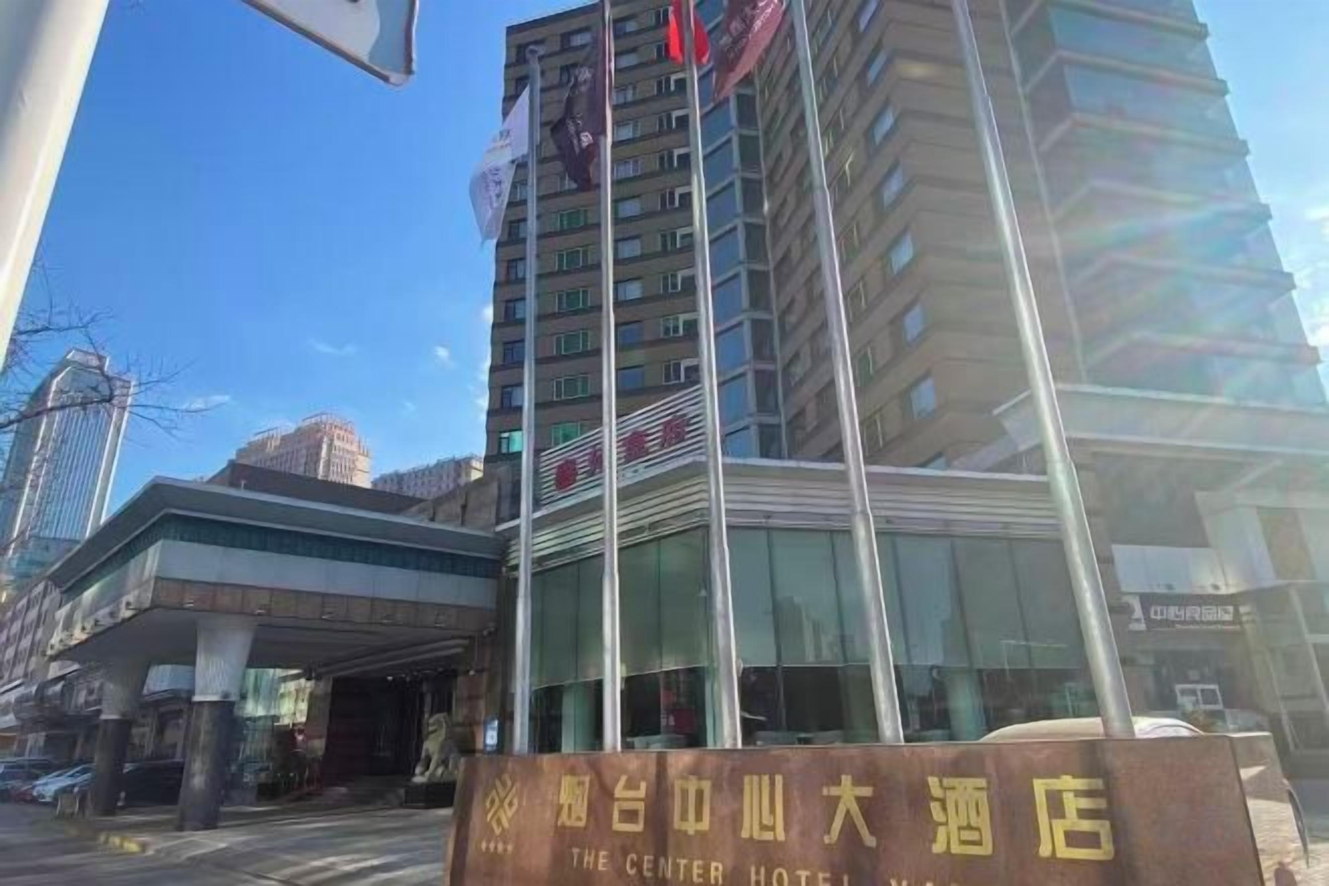 A hotel entrance with a sign in a foreign language, a glass facade, and a clear sky.