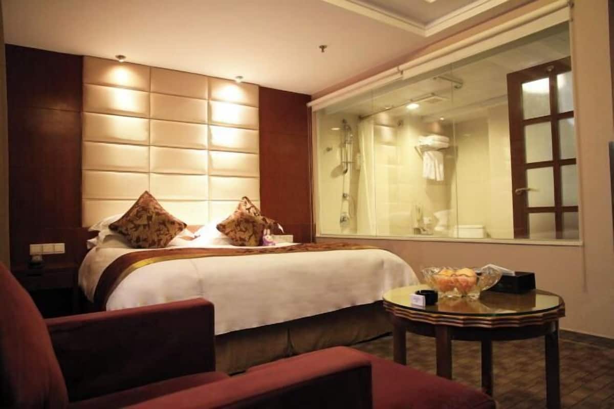 A hotel room with a bed, a round table with a glass top, a large window with a view of a bathroom, and a wooden headboard.
