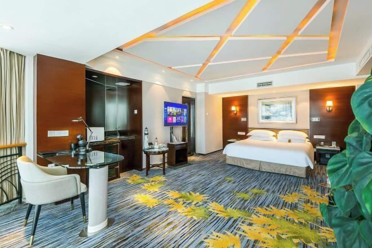 A hotel room with a bed, a desk, a TV, and a large plant.