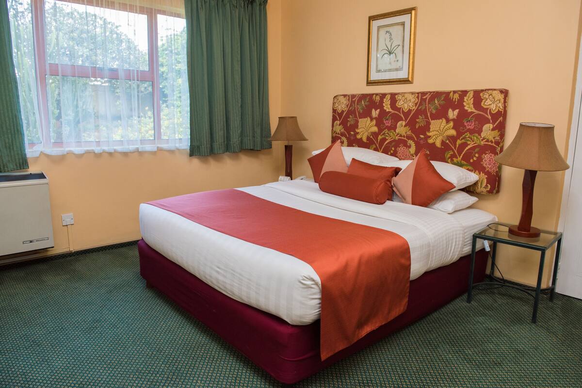A hotel room with a large bed, two bedside lamps, a window with curtains, and a radiator.
