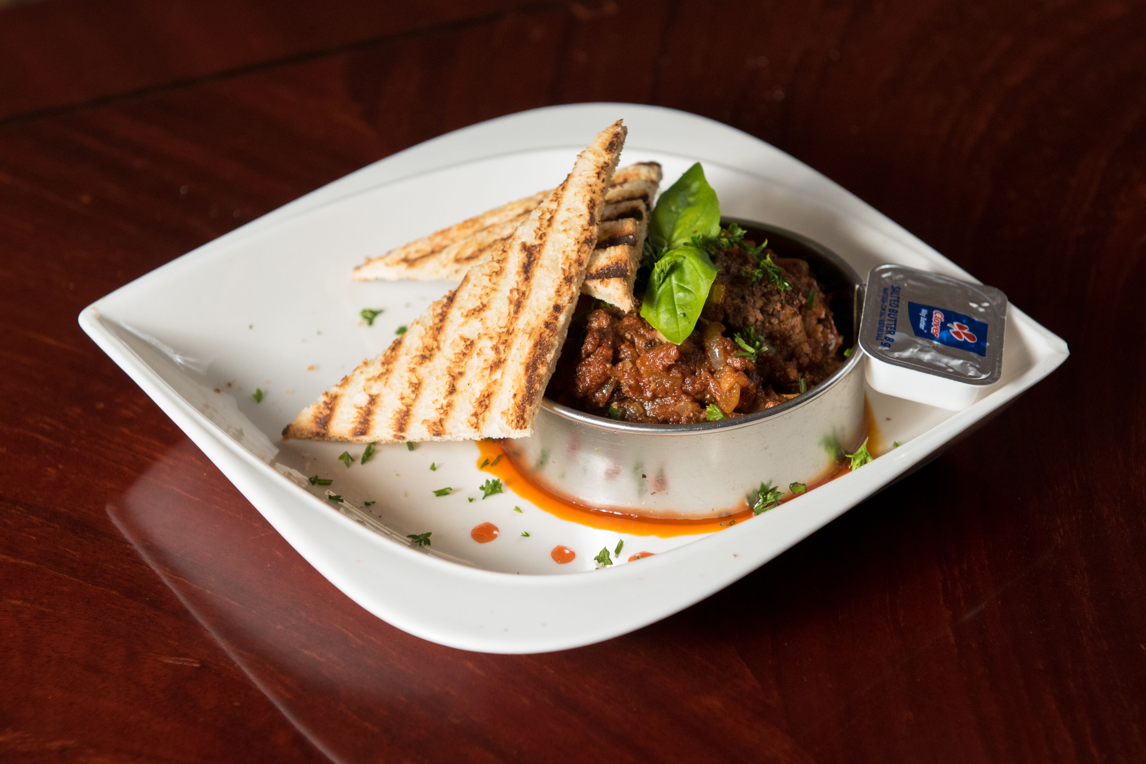 A square white plate with grilled bread, a bowl of meat dish, and a small container of sauce.