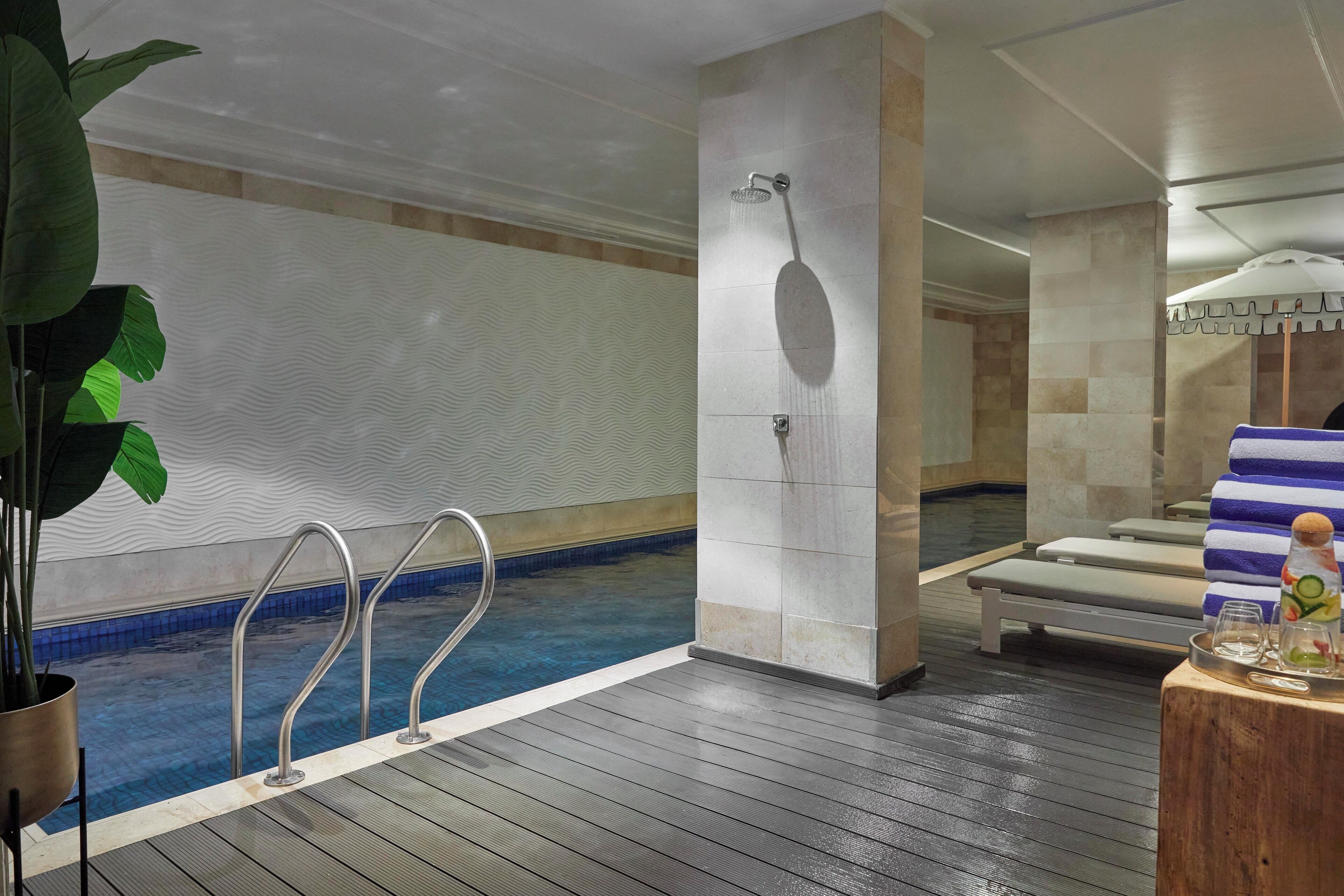Una piscina interior con plataforma de madera, zona de duchas y sillones de descanso con toallas.