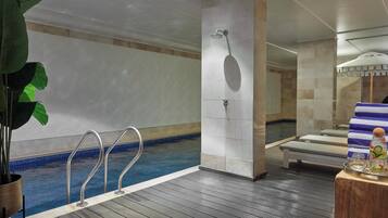 Una piscina interior con plataforma de madera, zona de duchas y sillones de descanso con toallas.
