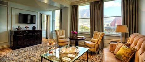 A well-appointed living room with a sofa, armchairs, a coffee table with a tray of pastries, a television, and a view of a mountainous landscape through large windows.