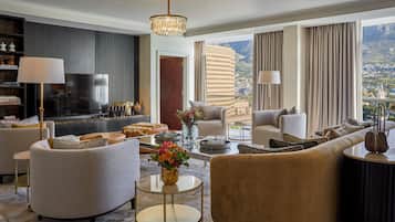 A modern hotel room with a large window offering a city view, a comfortable seating area with sofas and armchairs, a flat-screen TV, and a decorative vase with flowers.