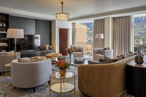 A modern hotel room with a large window offering a city view, a comfortable seating area with sofas and armchairs, a flat-screen TV, and a decorative vase with flowers.