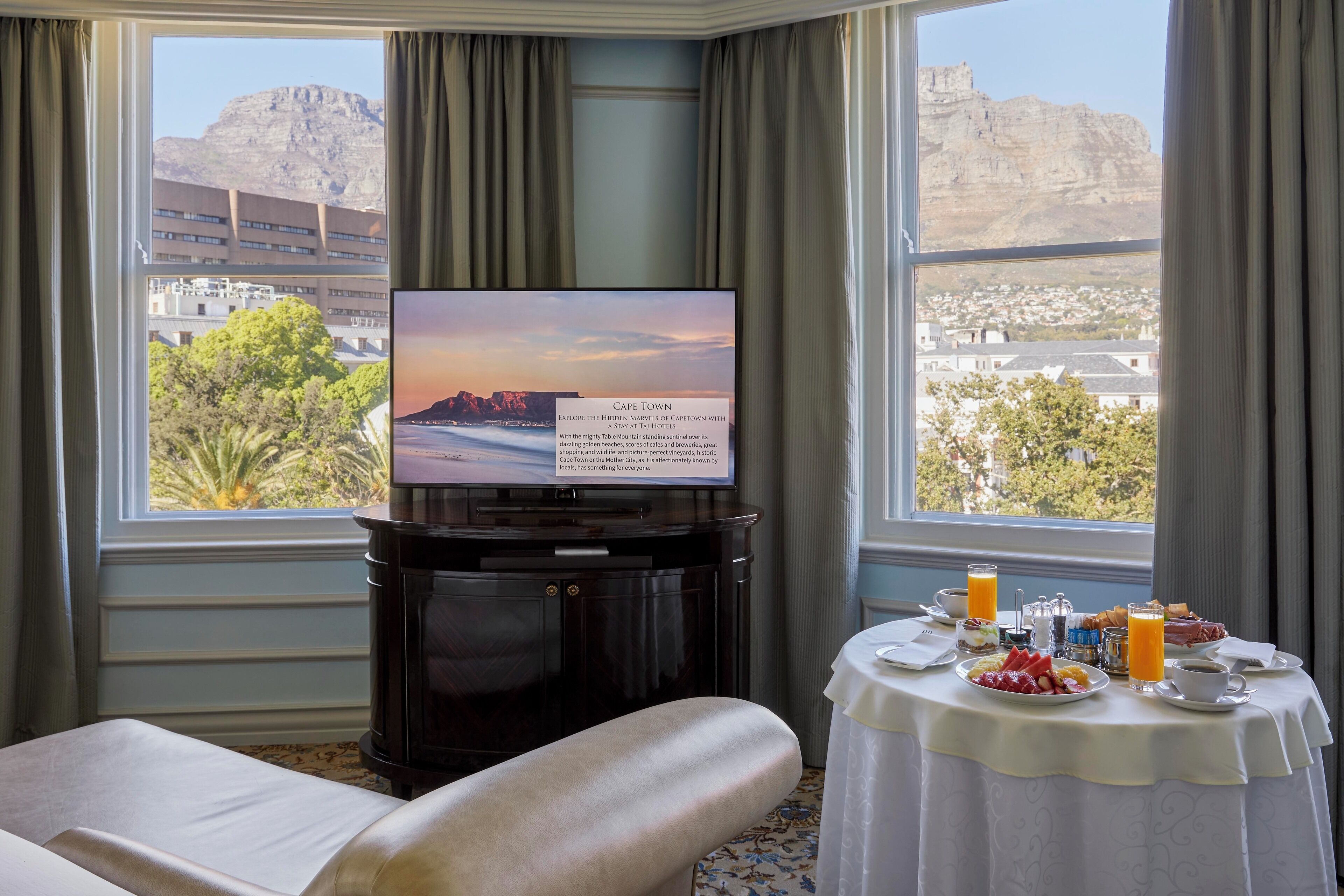 A hotel room with a large TV displaying a Cape Town view, a dining table with breakfast, and a comfortable seating area.