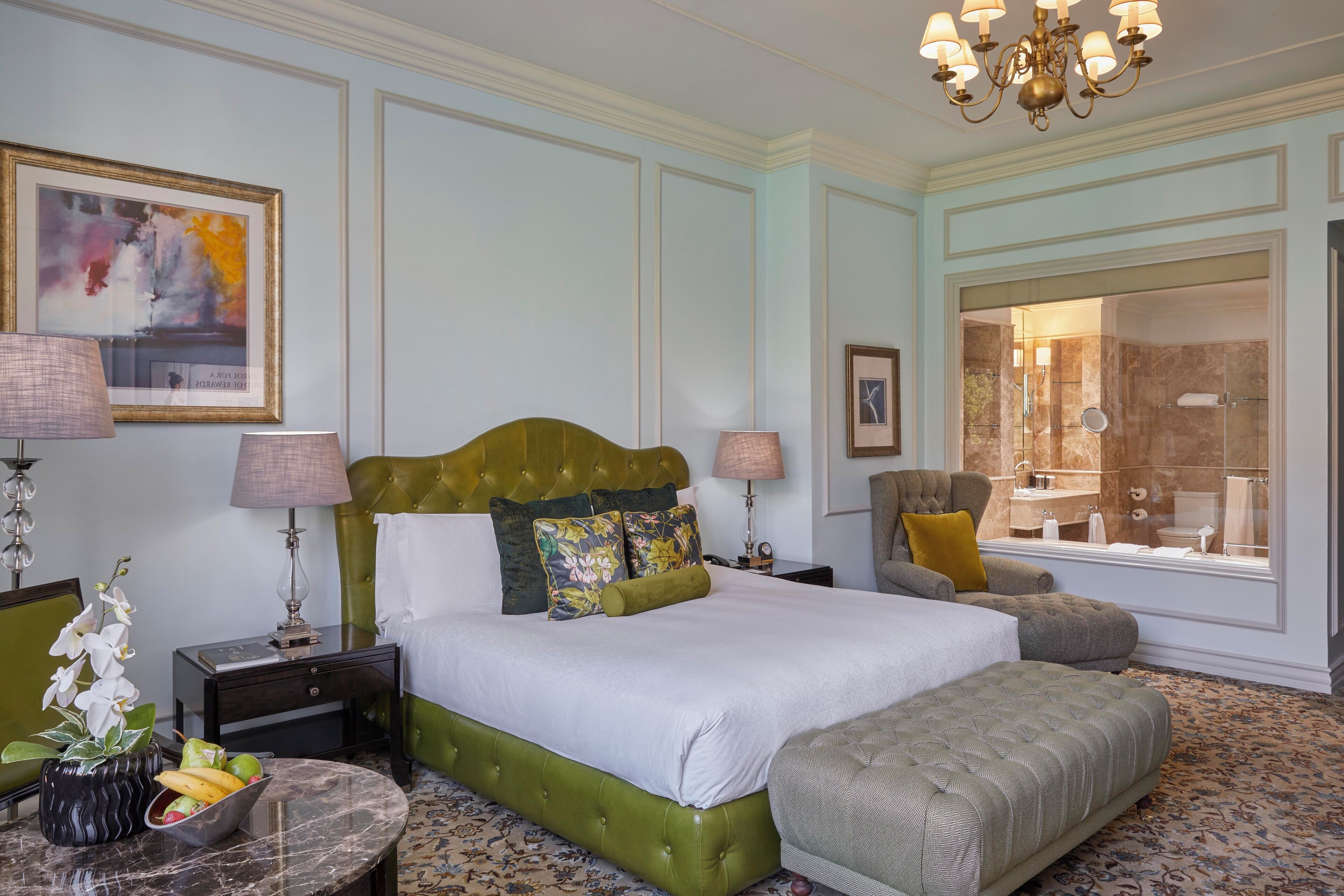 A spacious bedroom with a large bed, a sitting area, a bathroom with a glass shower, and a dining table with fruit.