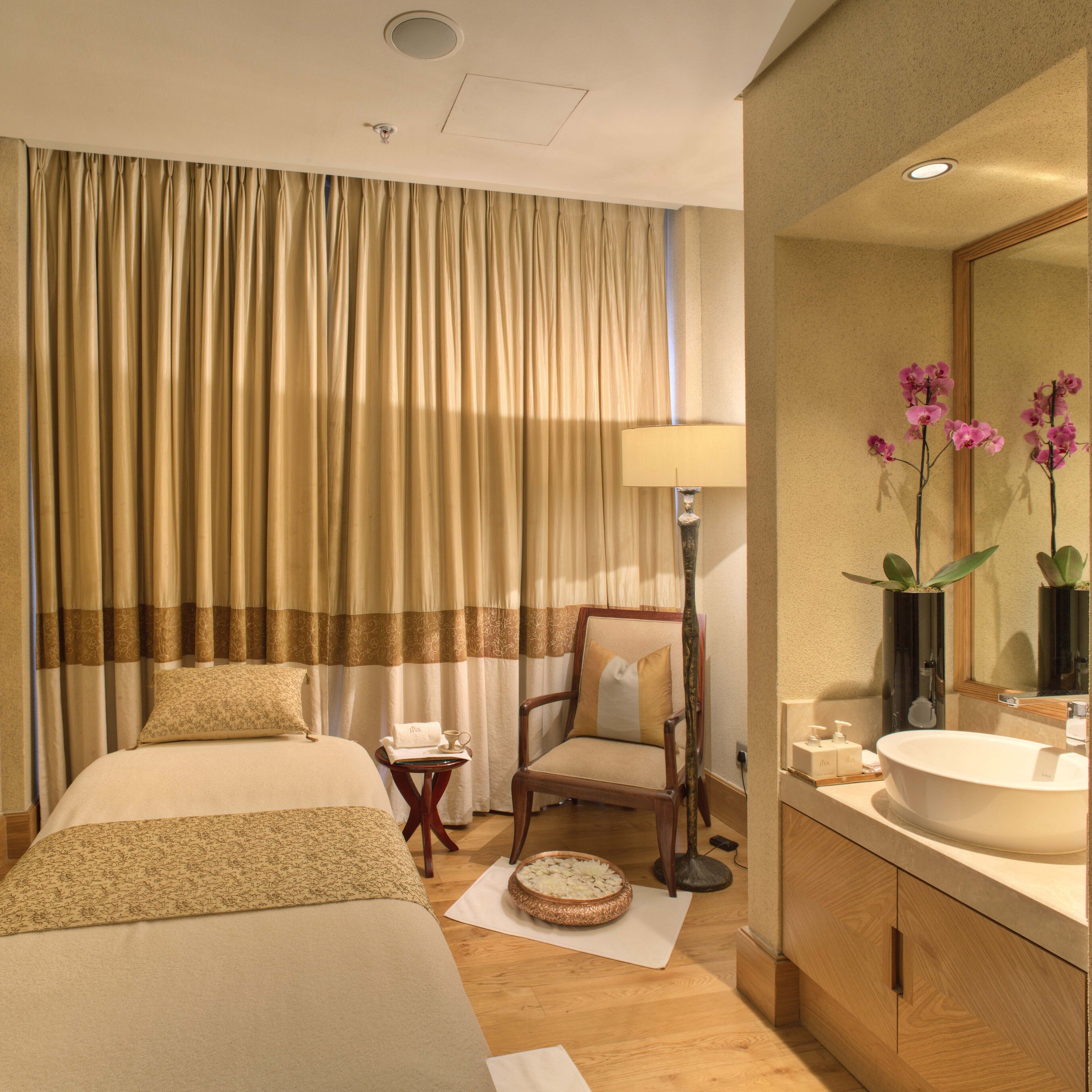 A hotel room with a bed, a chair, a bedside table with a lamp, a mirror, and a bathroom with a sink and mirror.
