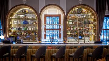 A bar area with three arched shelves filled with bottles, a bar counter with chairs, and a stained glass window.