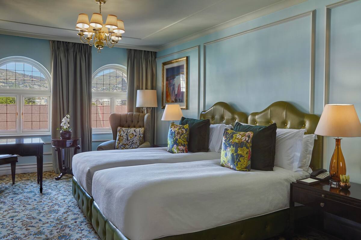 Une chambre d’hôtel avec deux lits, un lustre, un bureau et une vue sur la ville grâce à de grandes fenêtres.