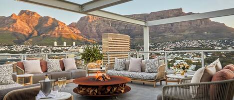 A rooftop terrace with a fire pit, seating area, and a view of mountains and buildings.