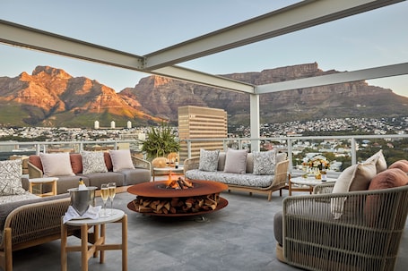 A rooftop terrace with a fire pit, seating area, and a view of mountains and buildings.