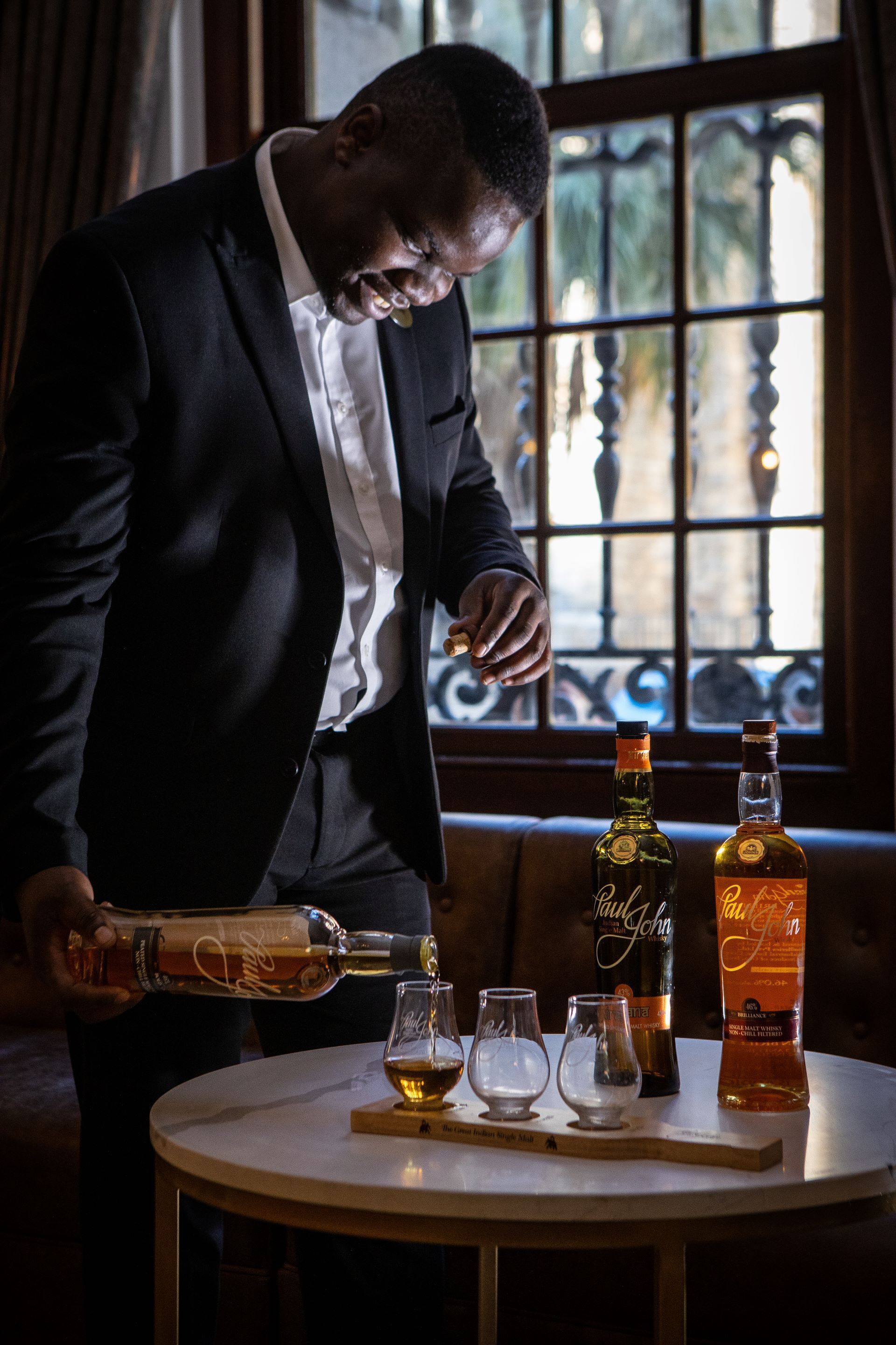 A man pouring a drink into a glass on a table with two bottles and two empty glasses.