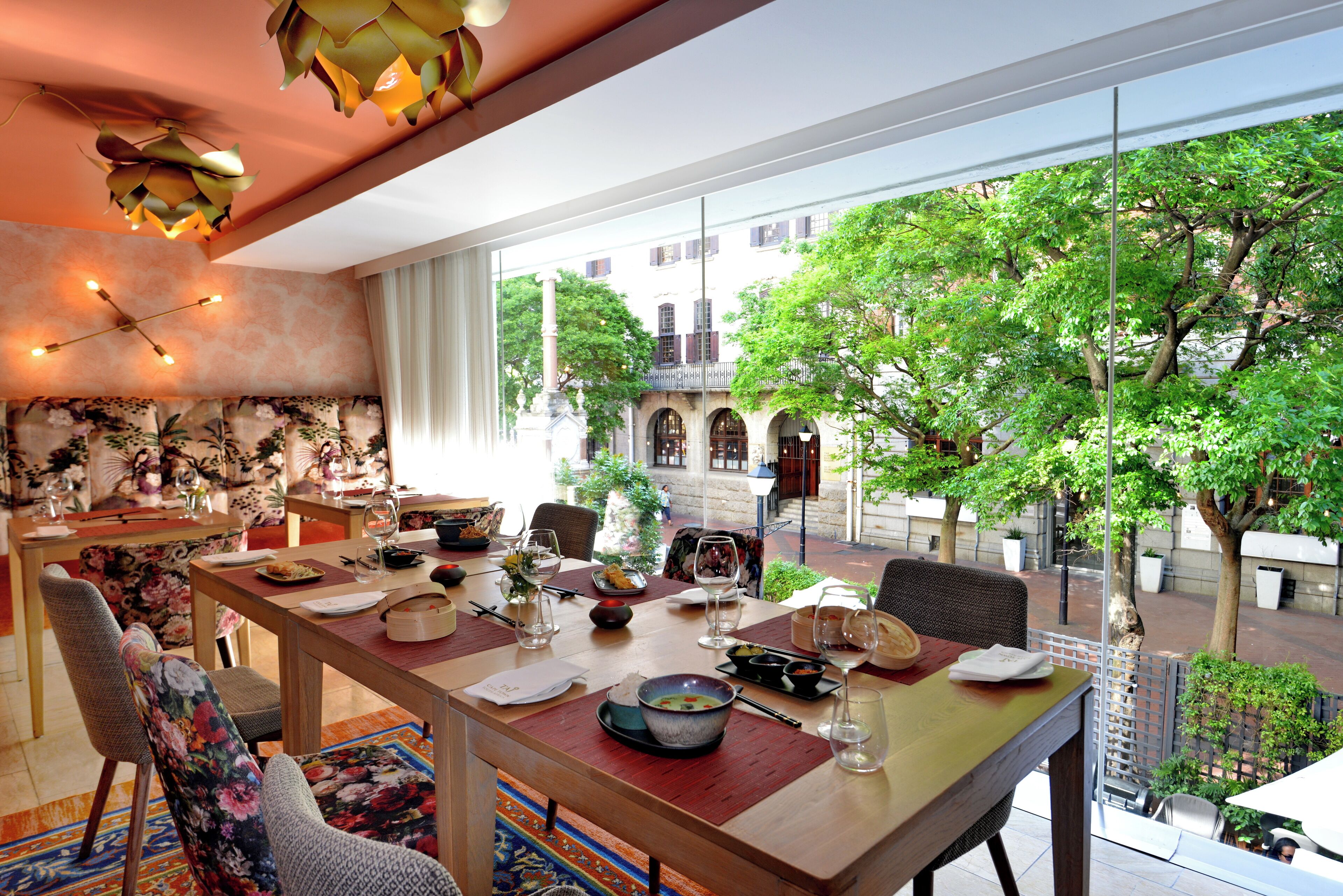 A dining area with a large table set for a meal, a view of a courtyard with trees and buildings, and a modern interior design.