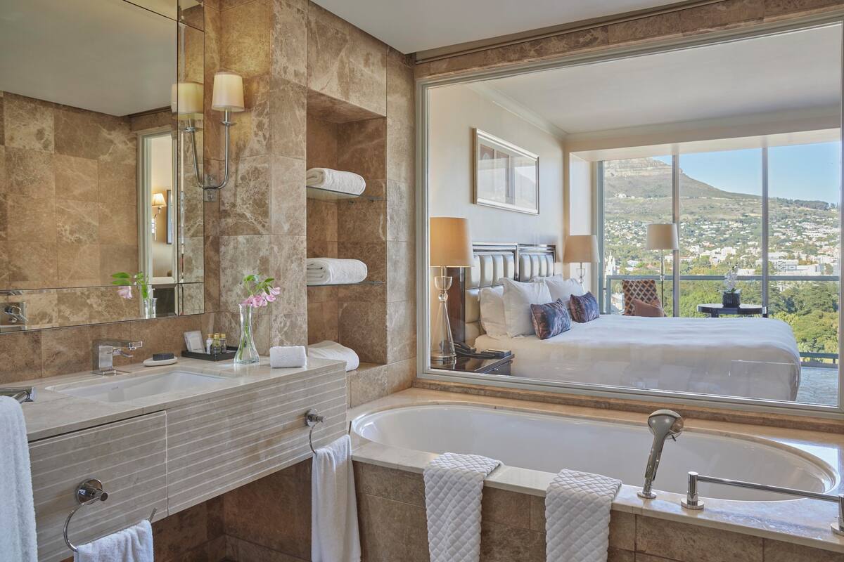 Une salle de bain moderne dotée d’une grande baignoire, d’un meuble-lavabo avec miroir et d’une grande fenêtre offrant une vue sur un lit et un paysage pittoresque.
