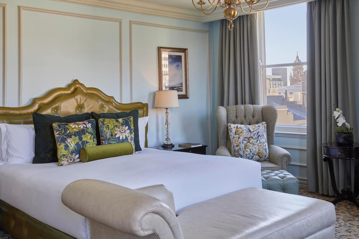 Une chambre d’hôtel avec un grand lit, une chaise, une lampe et une vue sur le paysage urbain par la fenêtre.