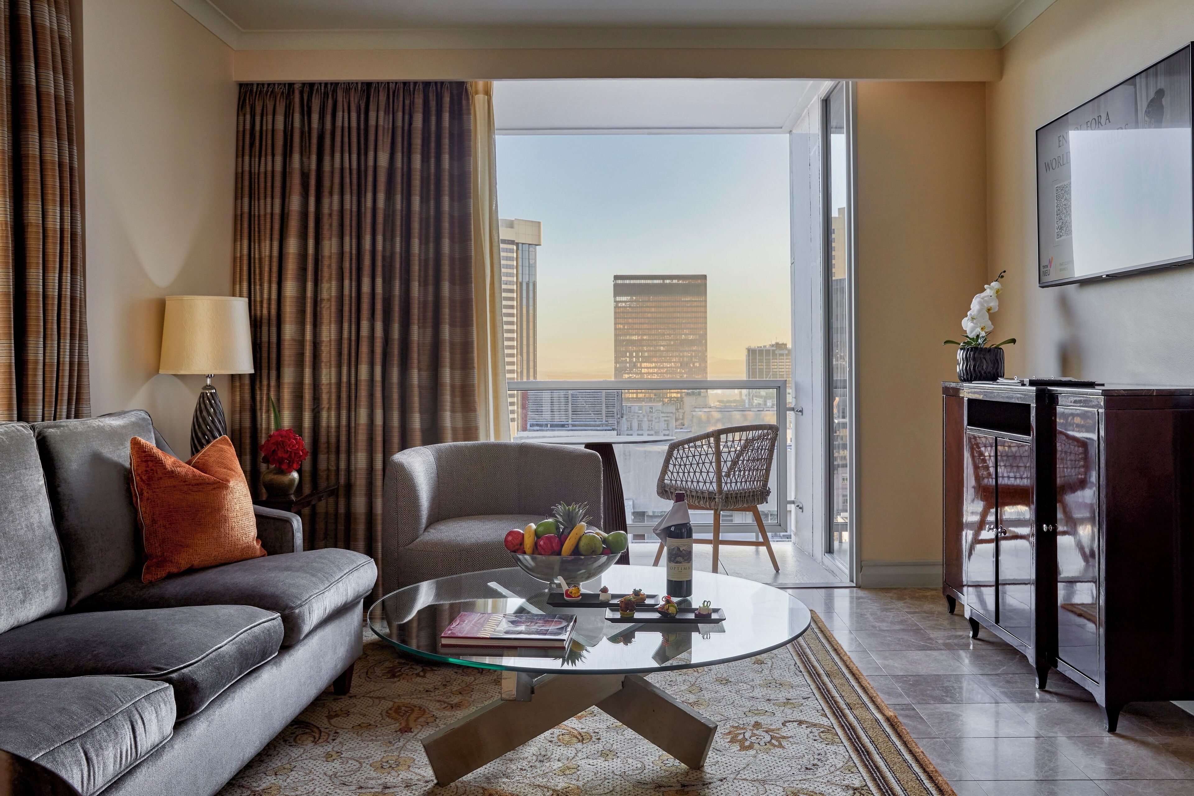 A modern living room with a sofa, a glass coffee table, and a view of a cityscape through large windows.
