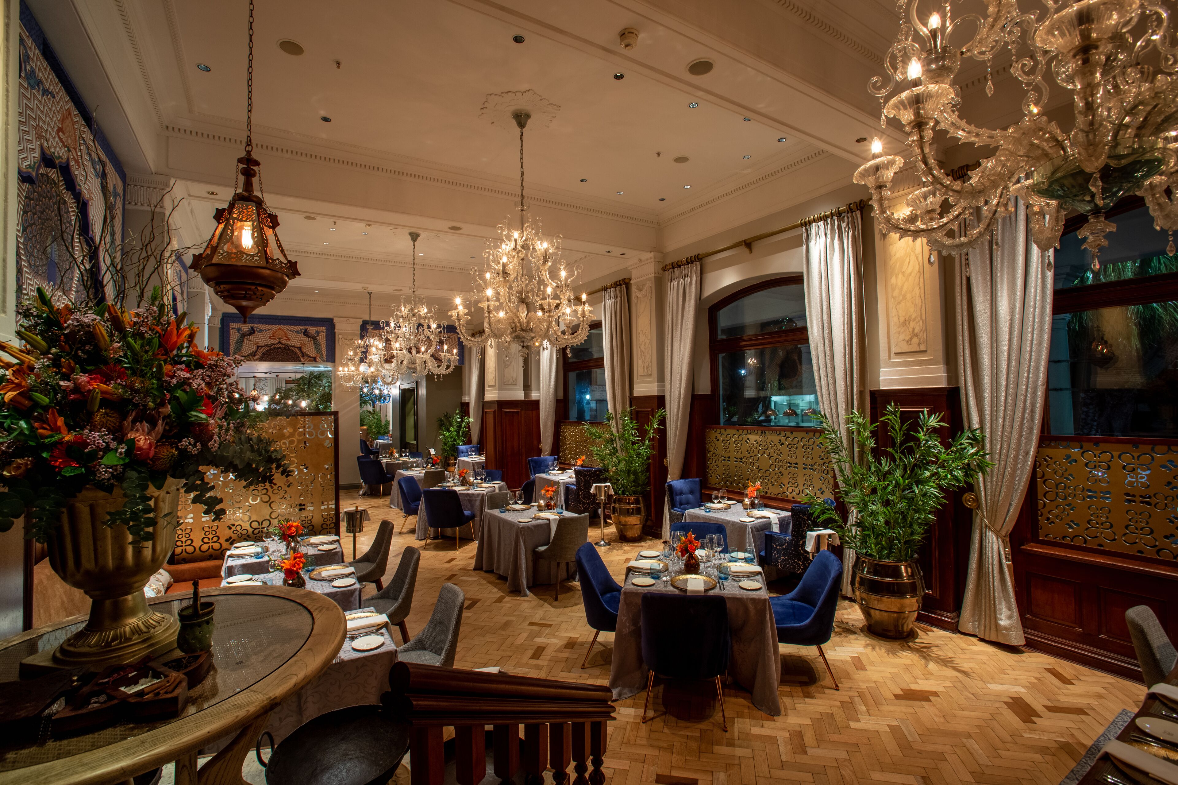 A spacious dining area with multiple tables and chairs, ornate chandeliers, and decorative plants.