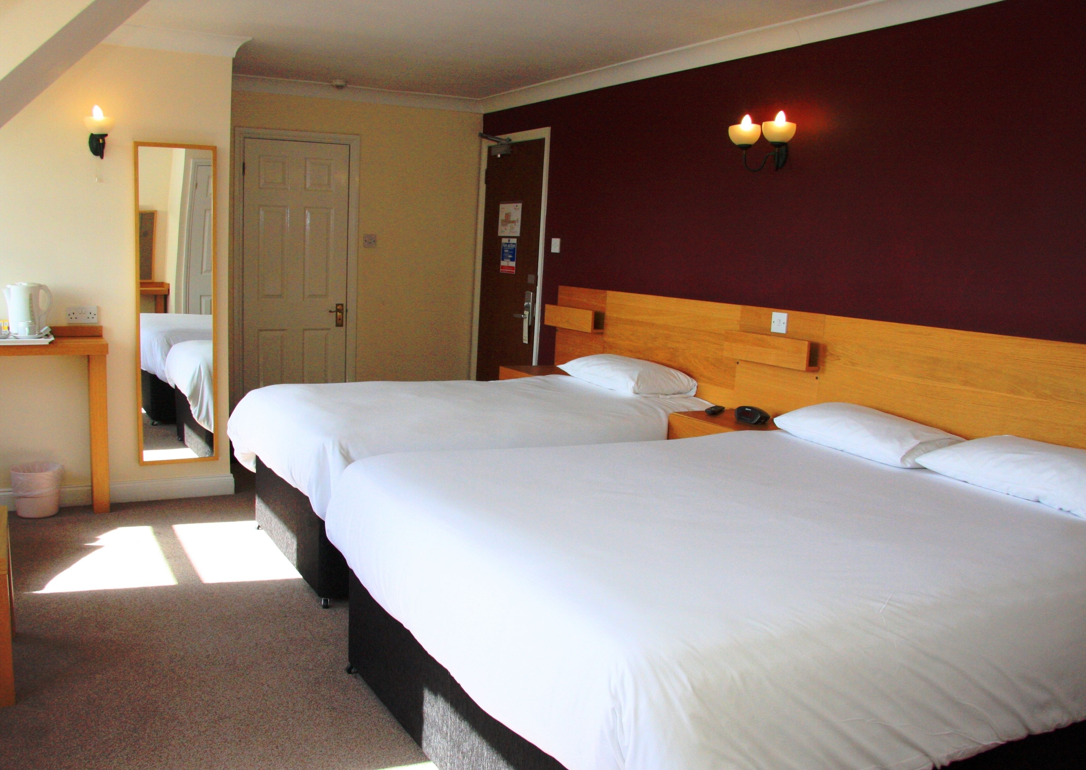 A hotel room with two beds, a wooden headboard, a mirror, a bedside table with a lamp, and a wall-mounted light fixture.