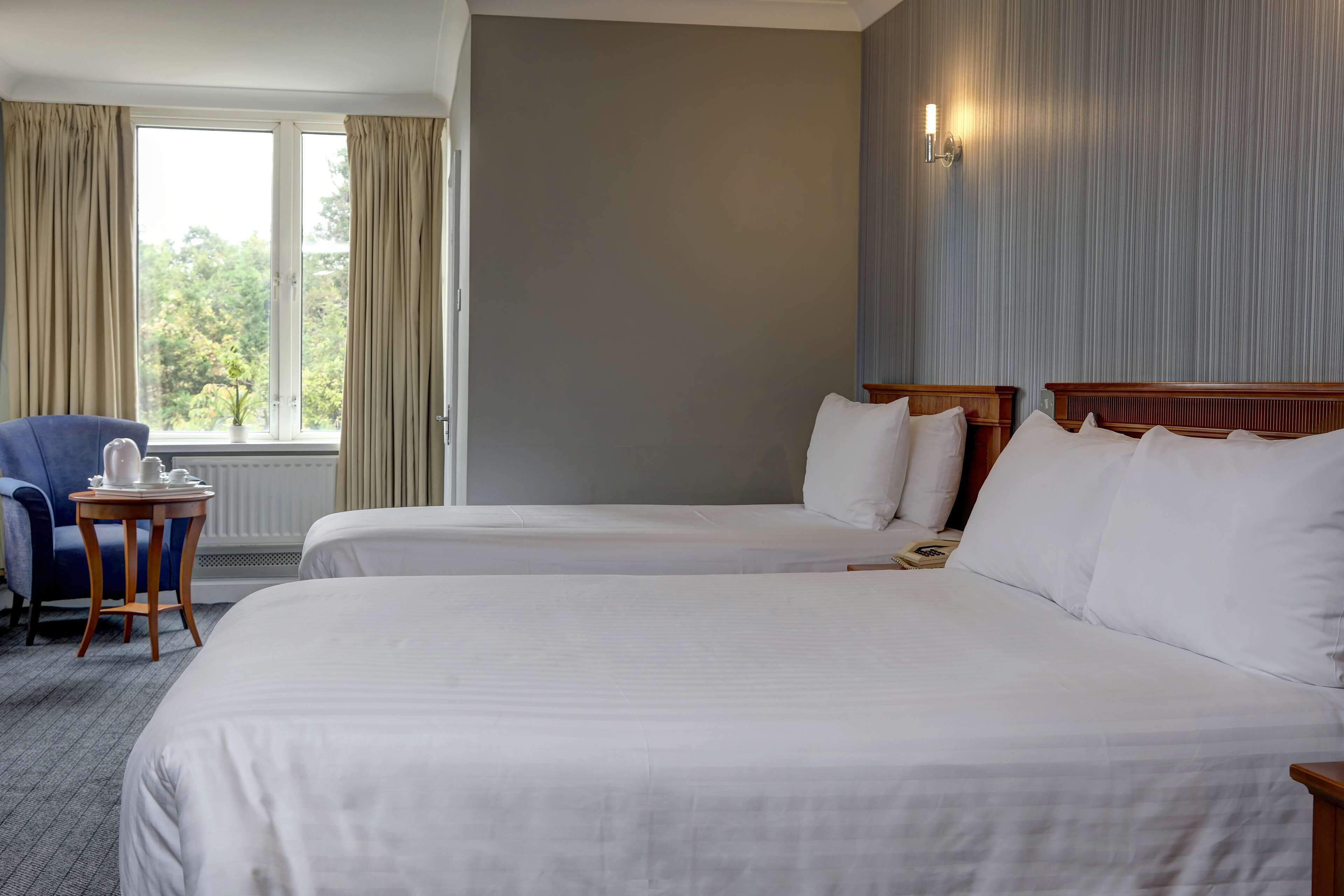 A hotel room with two beds, a small table with a kettle, a chair, and a window with curtains.