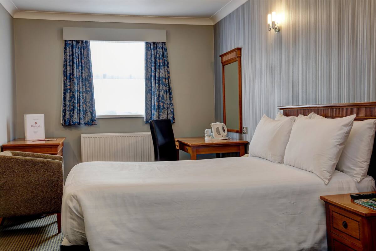 A hotel room with a large bed, a wooden nightstand, a chair, a table with a kettle, and a window with curtains.