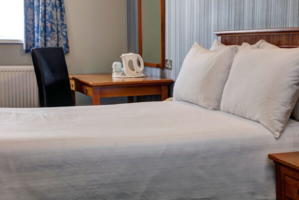 A neatly made bed with white linens, a wooden bedside table with a white kettle and cup, a chair, and a window with blue curtains.