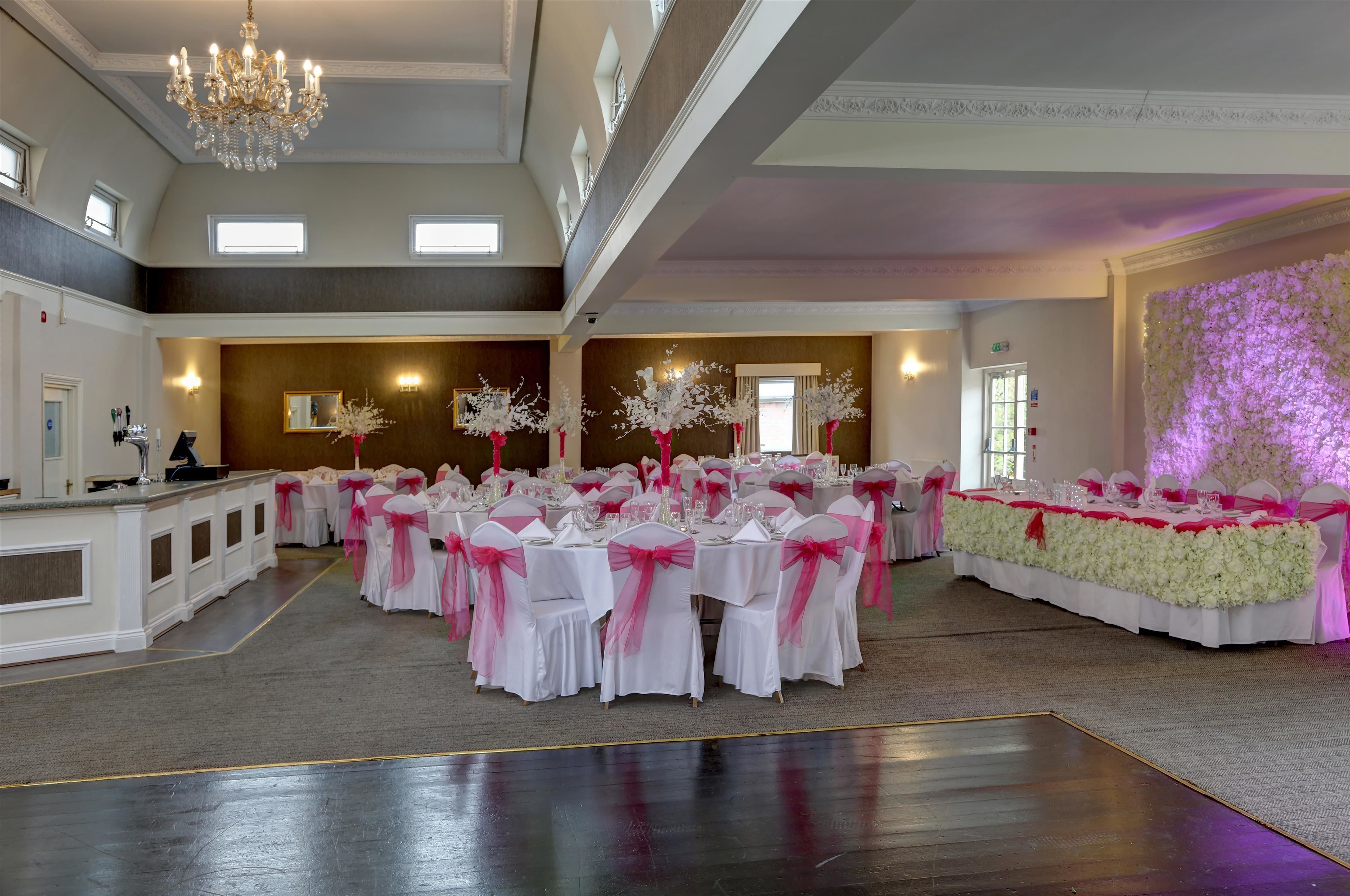Un amplio salón de banquetes con varias mesas redondas, cada una cubierta con manteles blancos y adornada con cintas rosas, preparado para un evento formal.