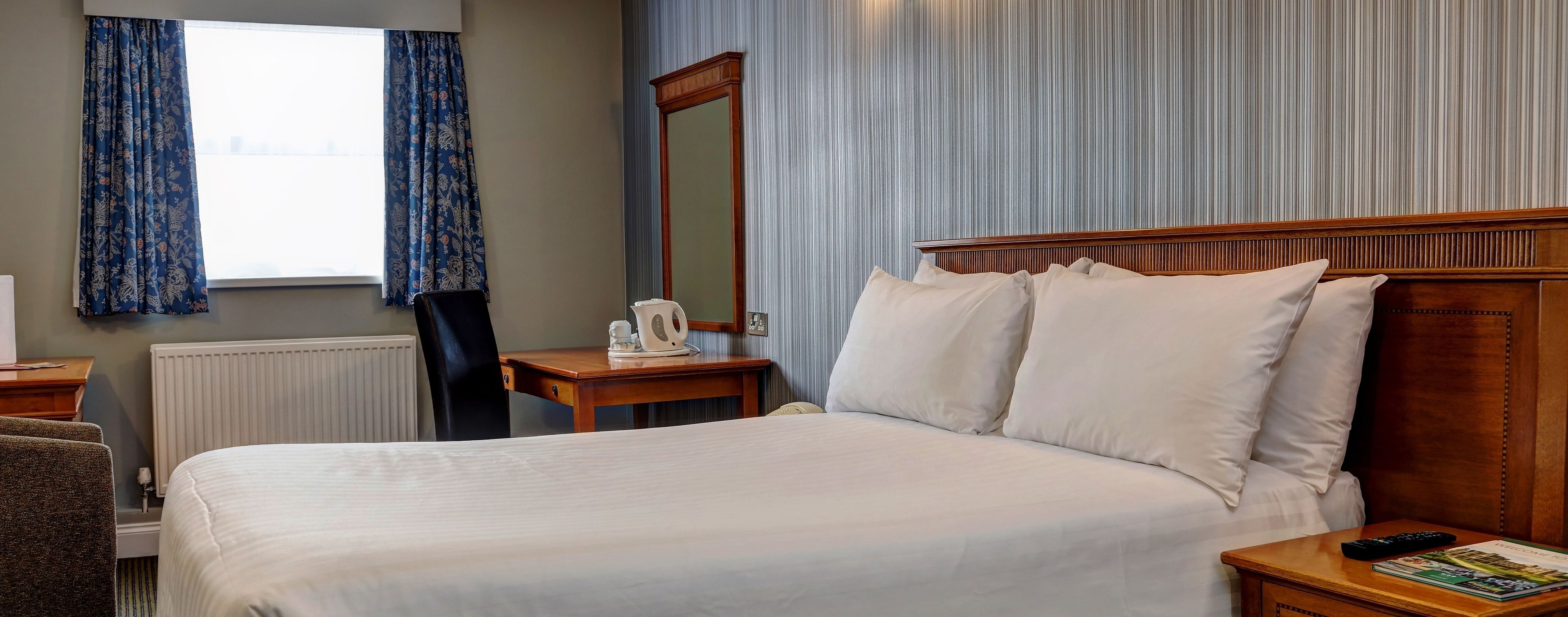 A hotel room with a large bed, a wooden headboard, a bedside table with a remote control, a mirror, and a window with curtains.
