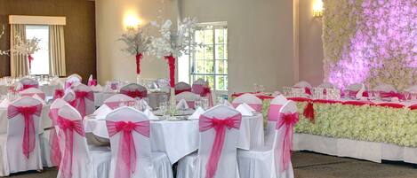 Un salón de banquetes con mesas redondas dispuestas para un evento formal, con manteles blancos y sillas adornadas con cintas rosas.