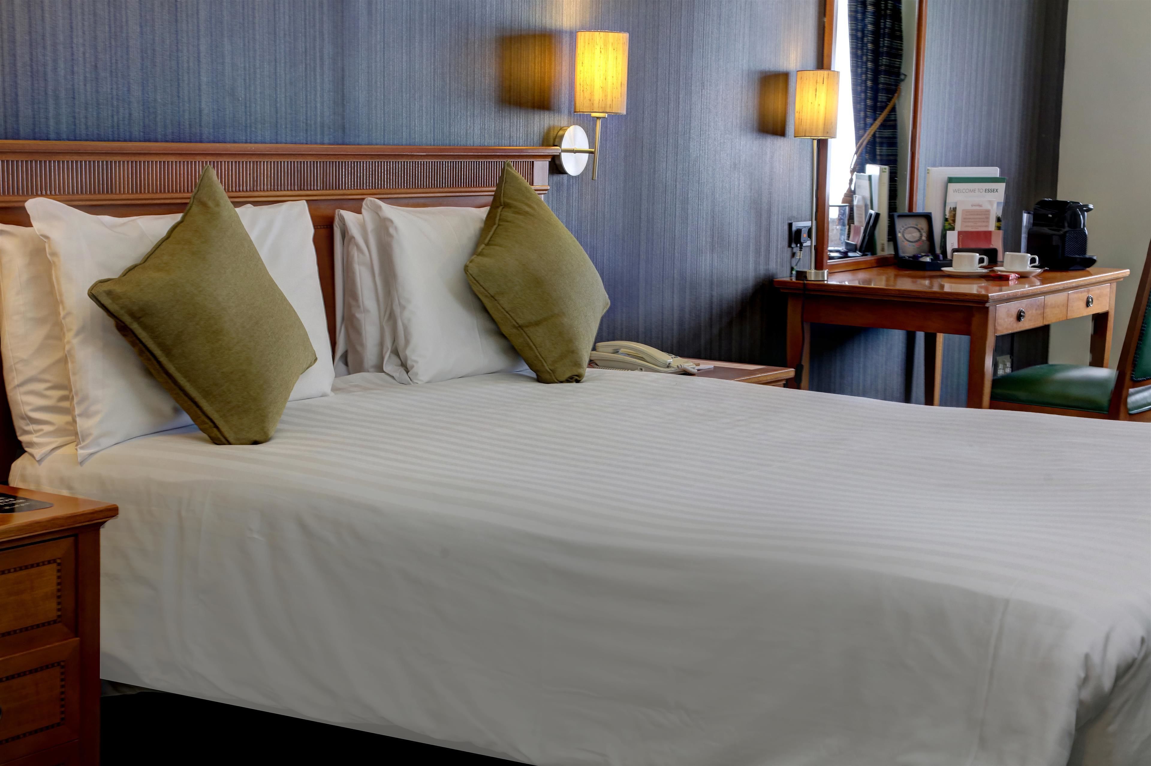 A hotel room with a large bed, two pillows, a wooden nightstand, a desk with a chair, and two wall-mounted lamps.