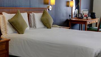 A hotel room with a large bed, two pillows, a wooden nightstand, a desk with a chair, and two wall-mounted lamps.