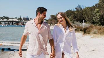 A couple walking on a sandy beach with a clear sky and a body of water in the background.
