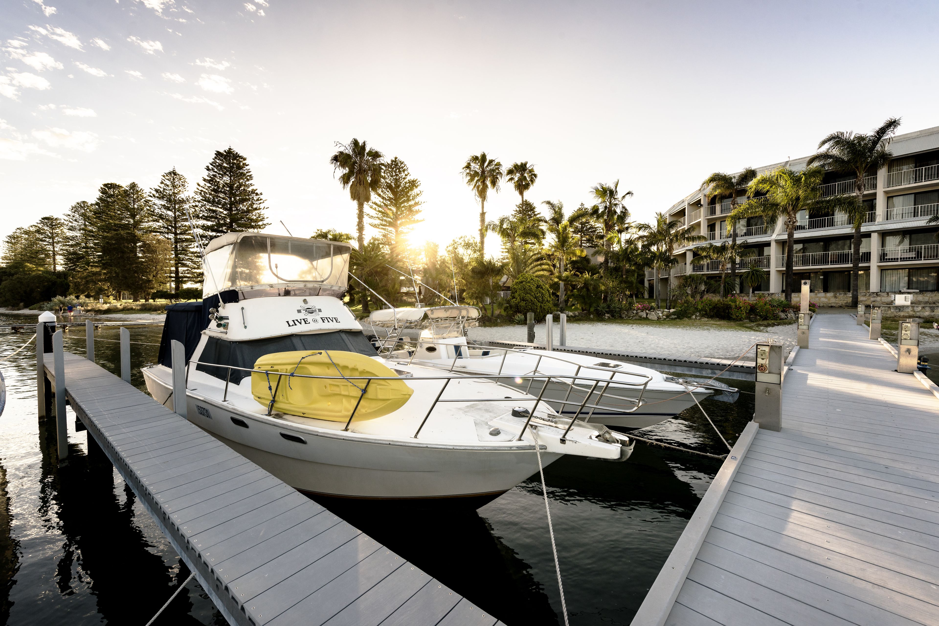 A marina with a white yacht docked at a wooden pier, surrounded by palm trees and a hotel in the background.
