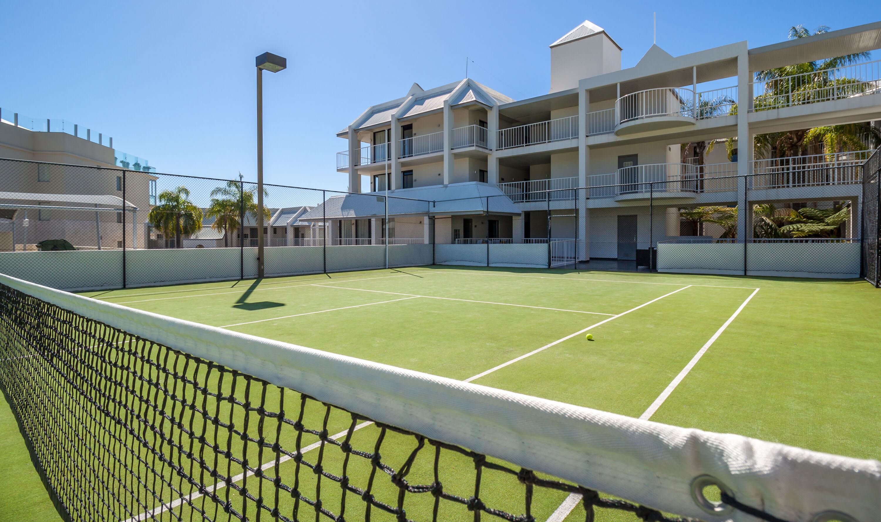 Una cancha de tenis con red, rodeada por una valla y un edificio con varios balcones.