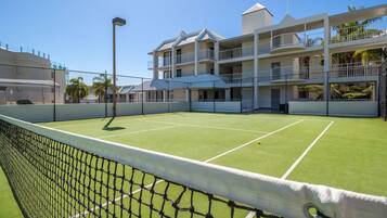 Una cancha de tenis con red, rodeada por una valla y un edificio con varios balcones.