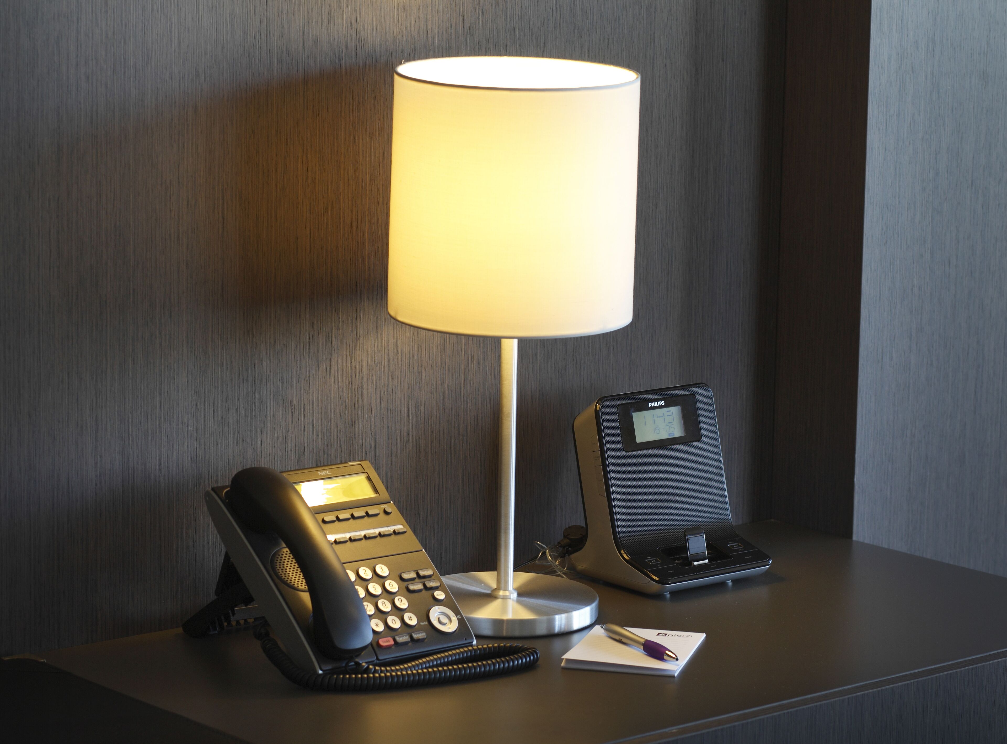 A hotel room with a desk featuring a desk lamp, a telephone, and a digital clock.