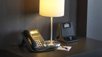 A hotel room with a desk featuring a desk lamp, a telephone, and a digital clock.