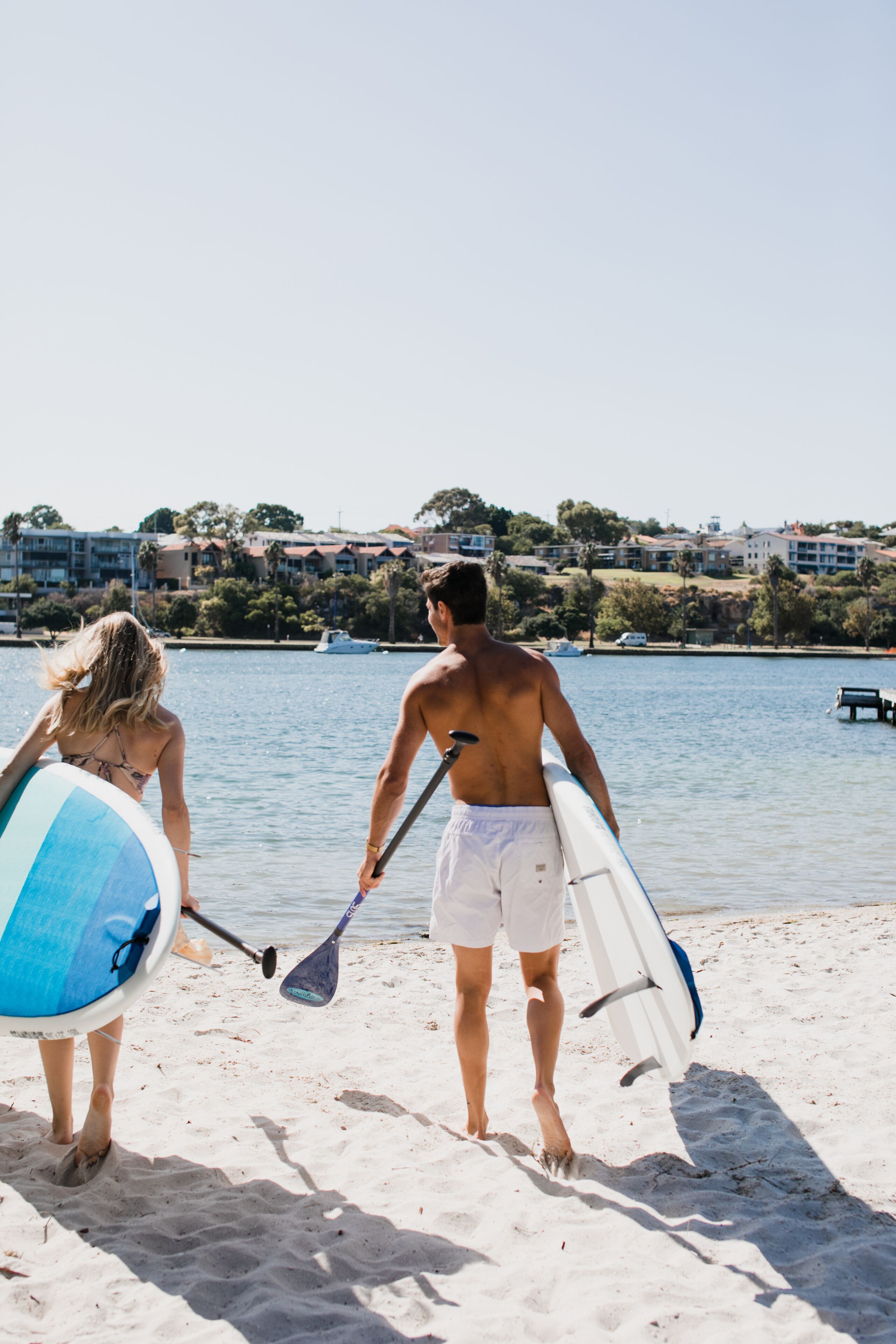 A beach scene with two people walking towards the water, each holding a paddle and a surfboard.