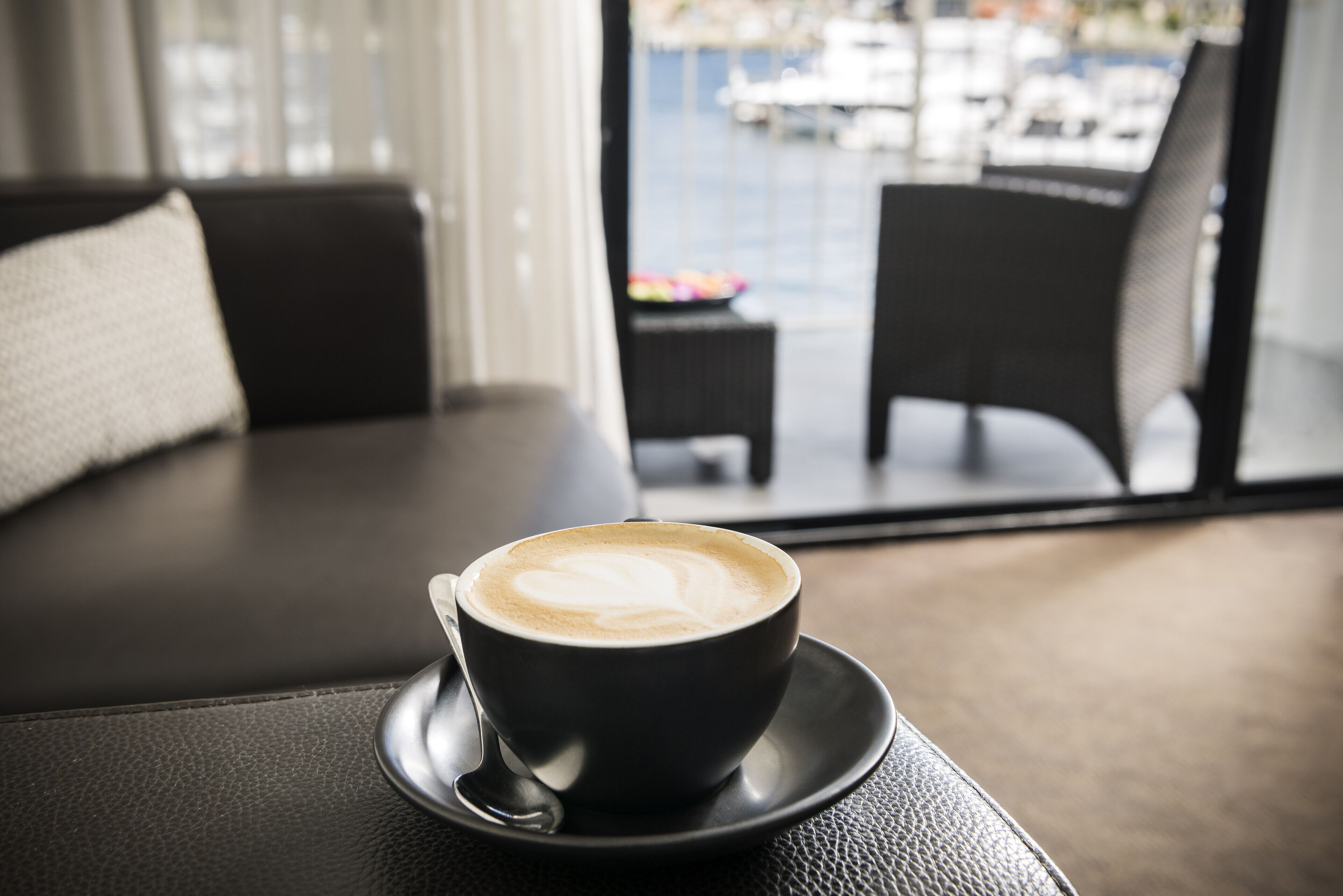 Une tasse de café posée sur une soucoupe, sur une table, avec en arrière-plan une vue sur un balcon et un espace extérieur aménagé avec des sièges.